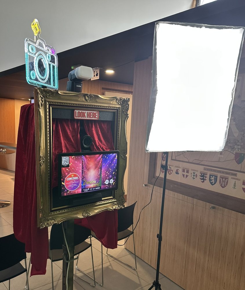 Photomaton installé en salle : cadre doré,
             néon en forme d'appareil, rideaux de velours, écran aux couleurs de l'événement
             et éclairage de studio